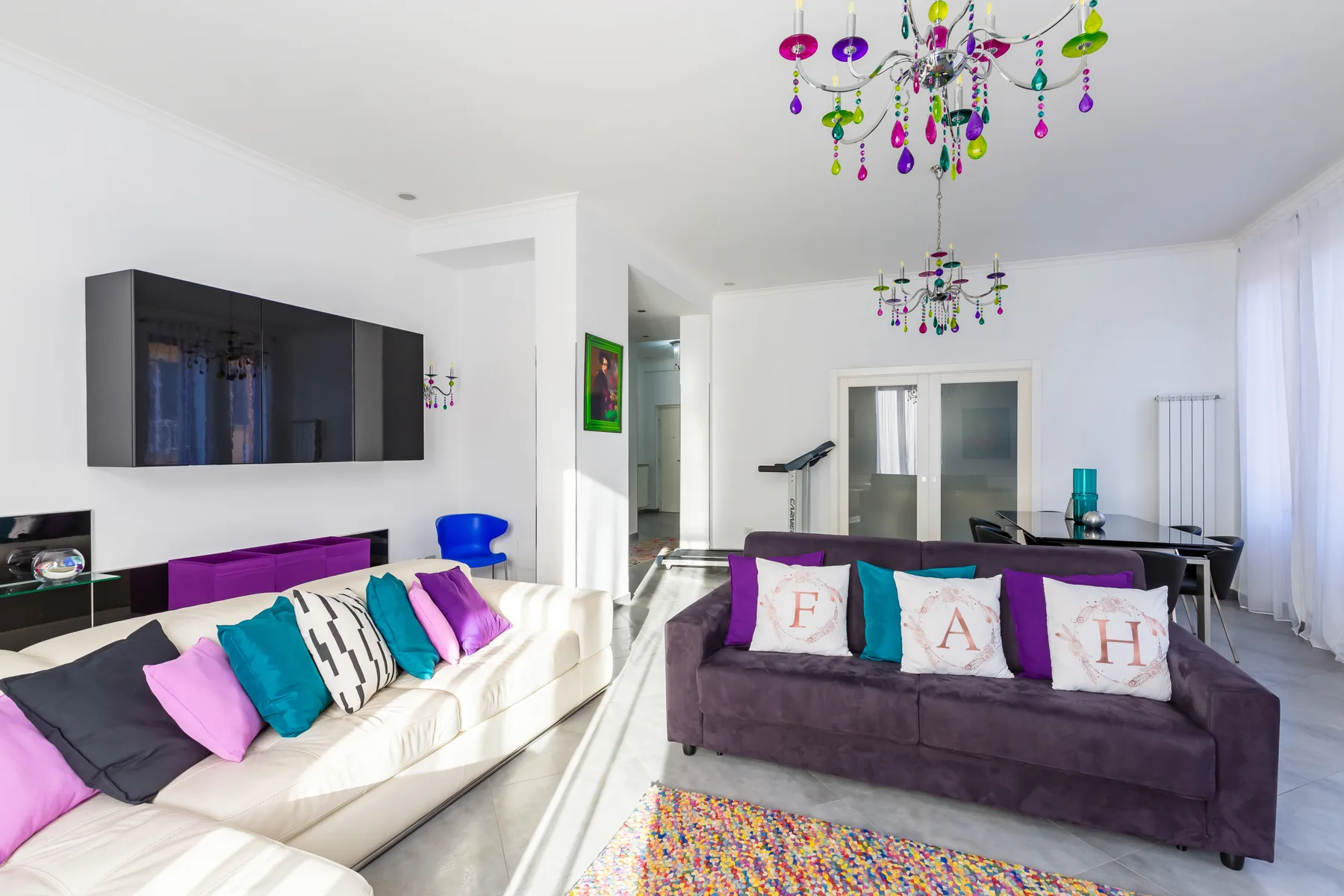 A bright living room with seating, a wall-mounted television, and colorful hanging lights.