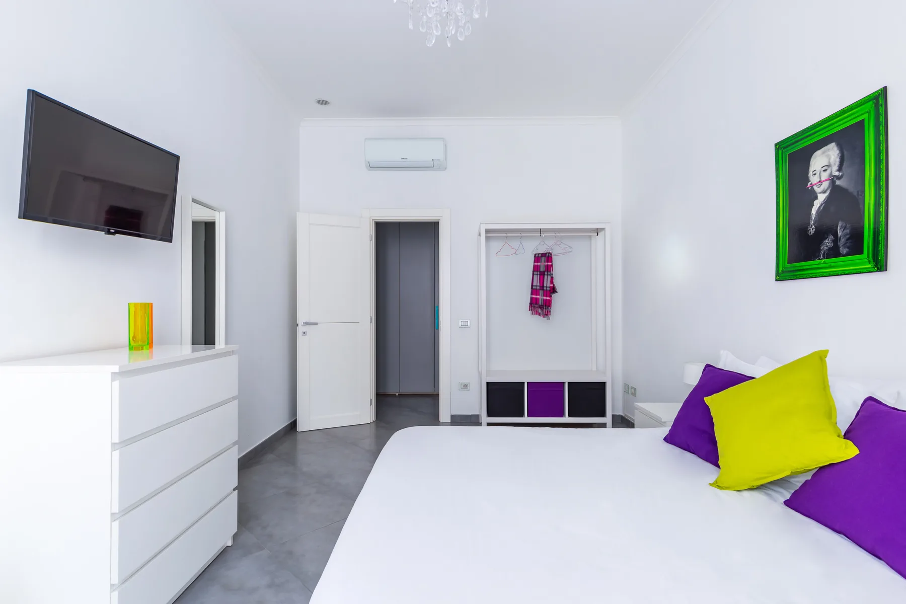 A bright white bedroom with a bed, wall-mounted television, and colorful pillows.
