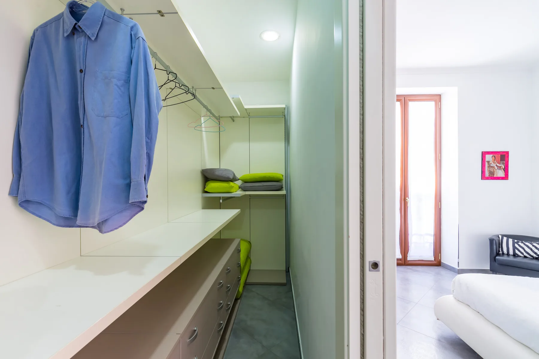 A dressing area with hanging clothes and a view into a bright bedroom.