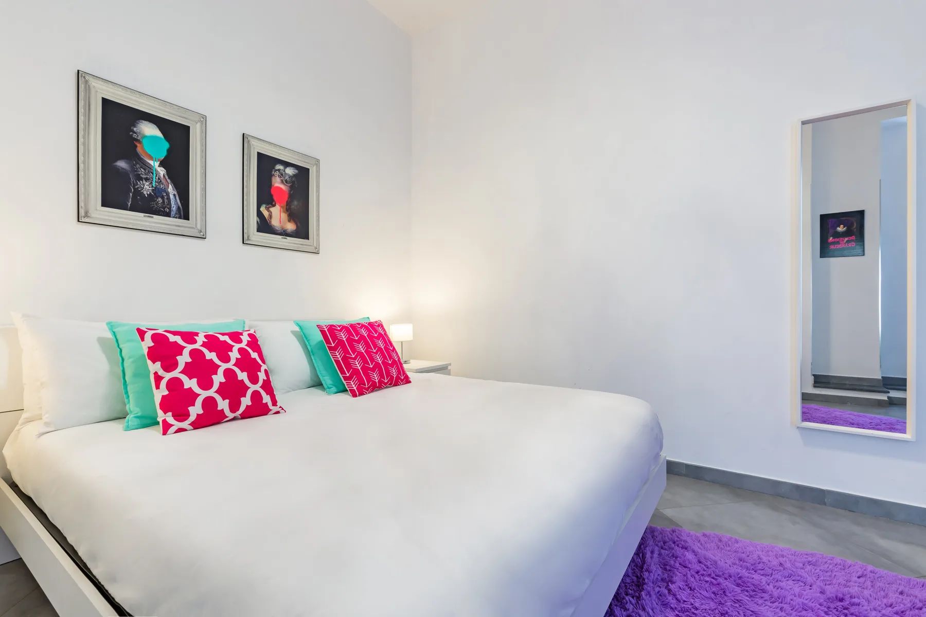 A bright bedroom features a large bed with colorful pillows and framed artwork.