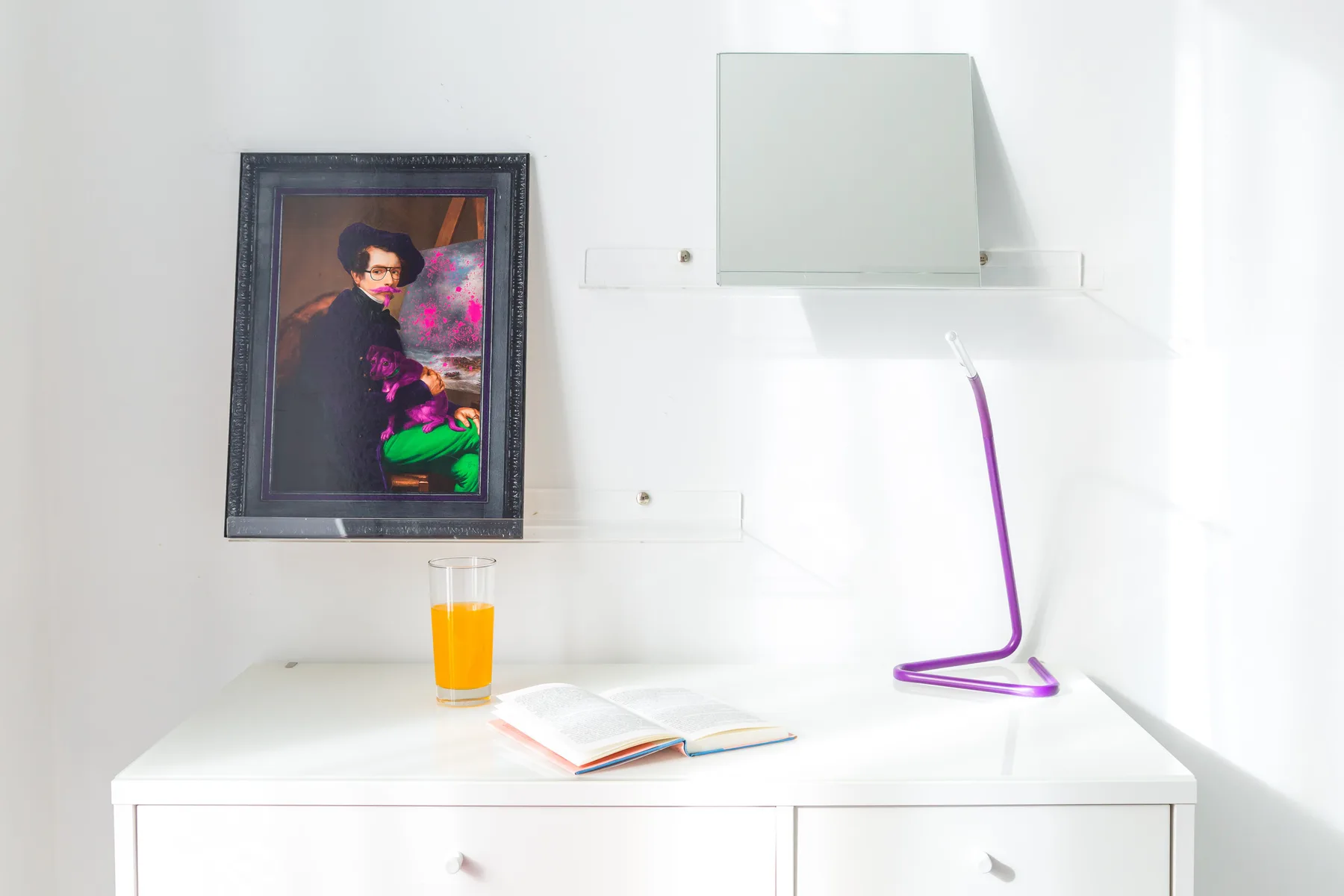 A bright white desk with a framed artwork, an open book, and a purple lamp.