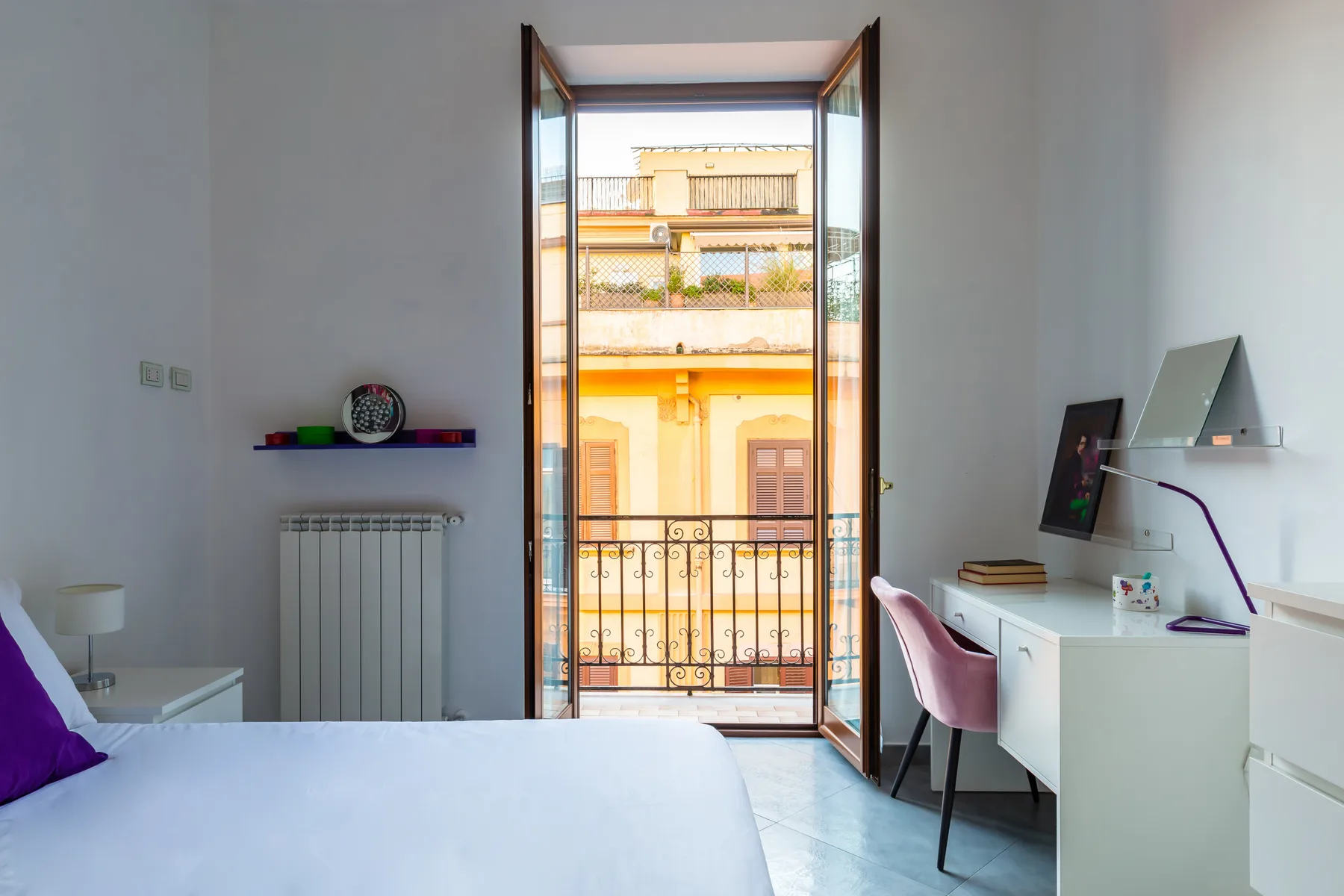 A bright bedroom with a bed, desk, and open balcony doors.