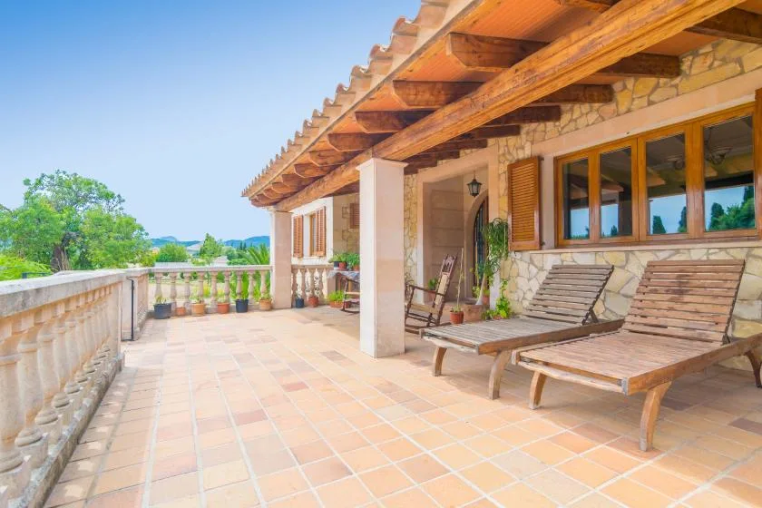 A sunlit tiled terrace with wooden loungers beside a stone-faced house.