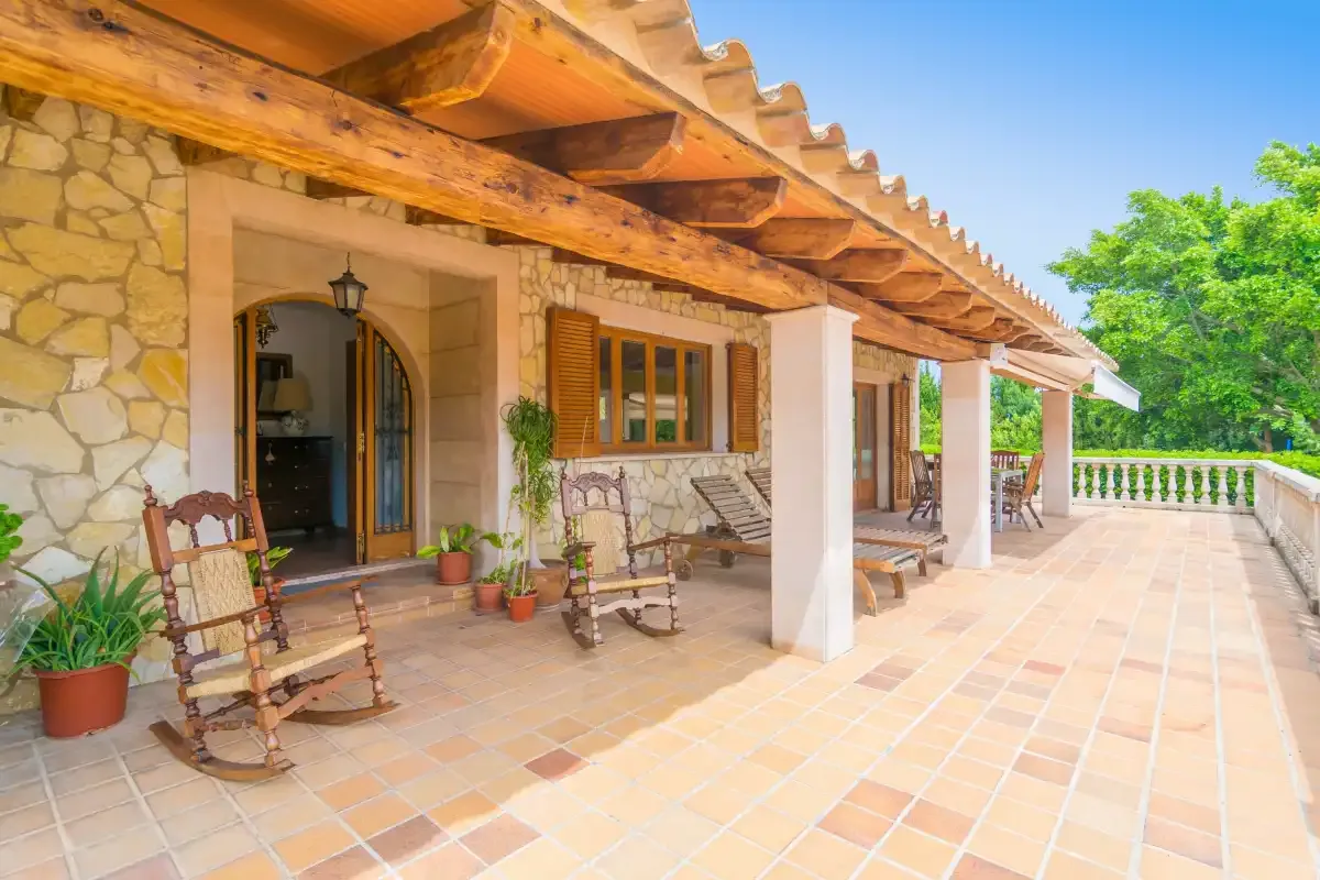 A stone house opens onto a covered terrace with seating and potted plants.