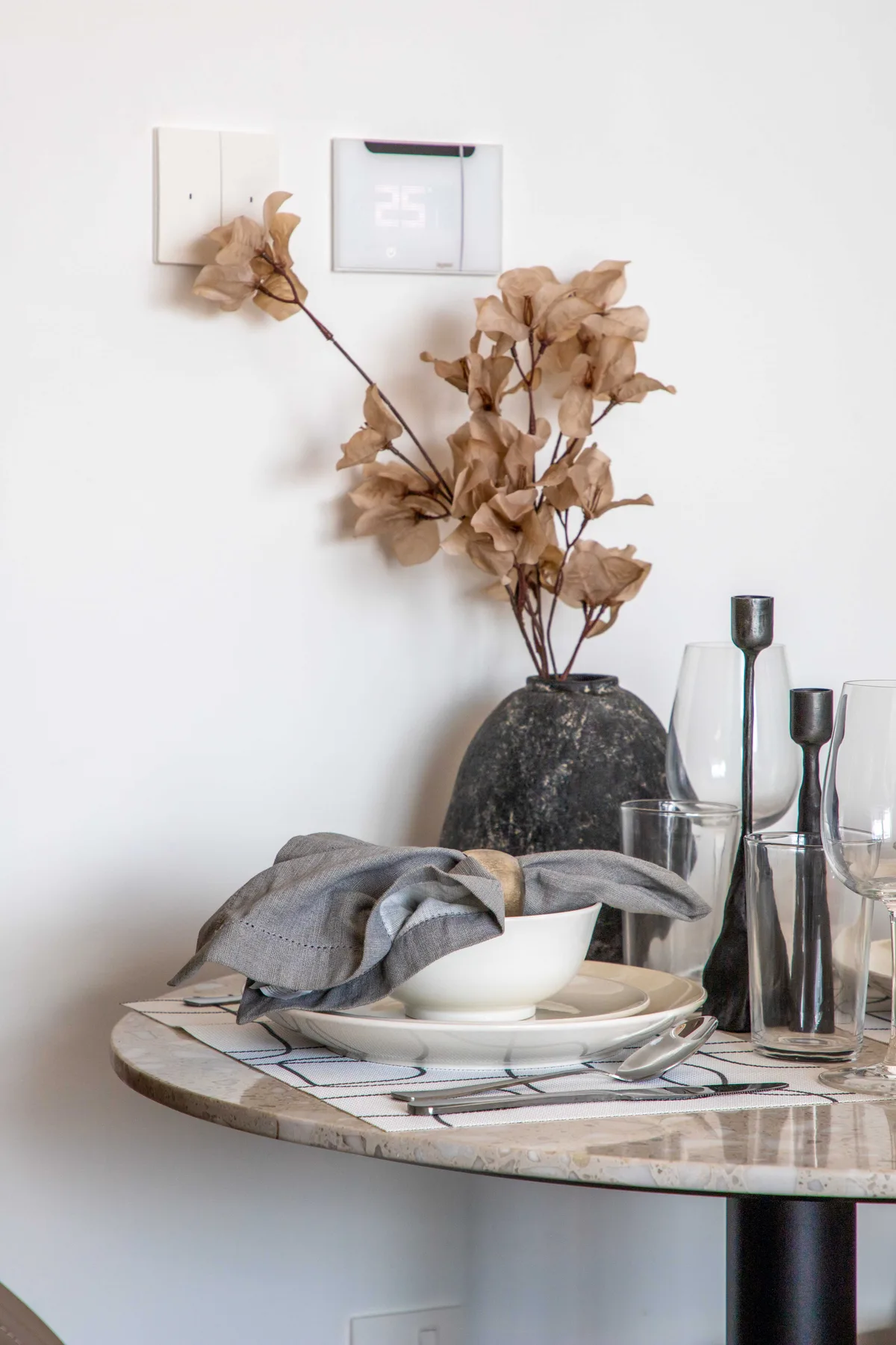 Una mesa de comedor con platos y vasos está bajo un jarrón con flores secas.