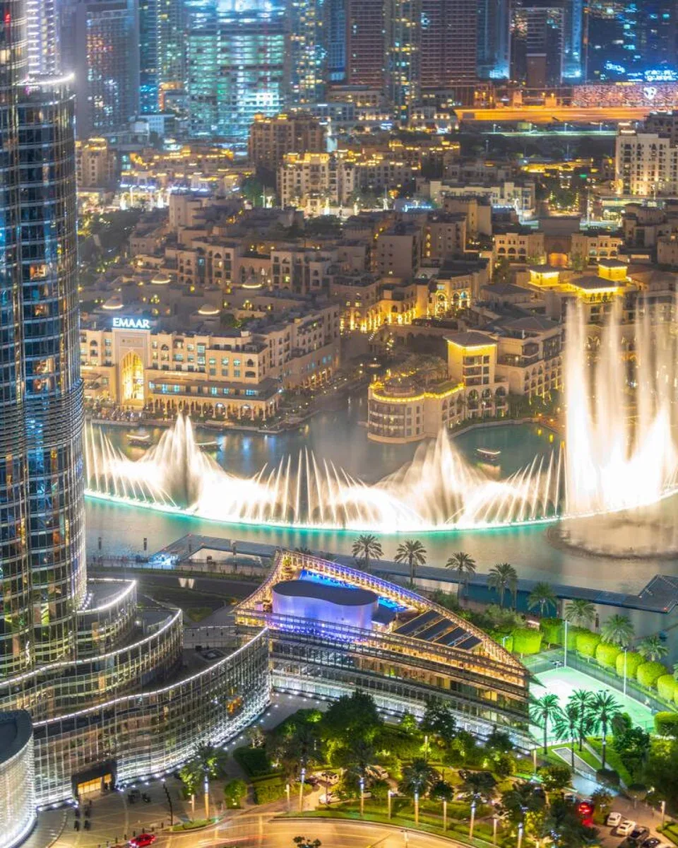A nighttime city skyline overlooks a brightly illuminated fountain show and surrounding buildings.