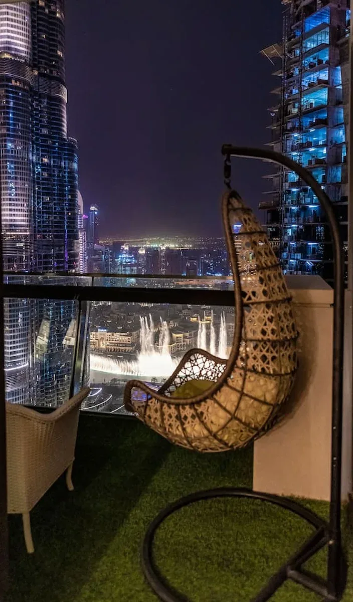 A balcony at night overlooks a brightly lit city skyline from beside a hanging seat.
