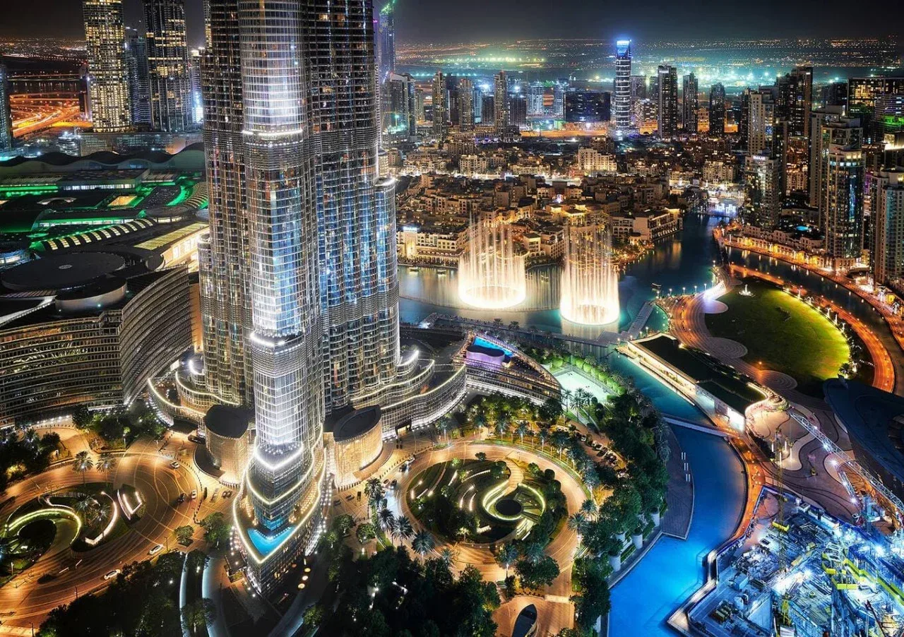 Nighttime cityscape with illuminated skyscrapers, fountains, and a curved pool.