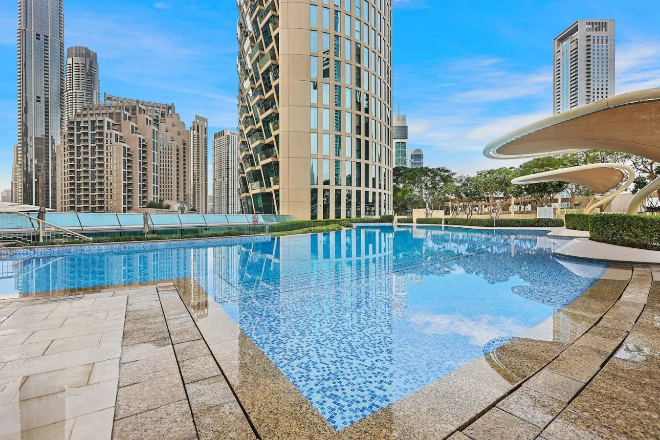 A swimming pool reflects nearby high-rise buildings and curved shade canopies.