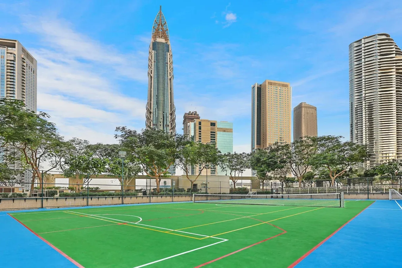 An outdoor sports court with green playing surfaces sits beneath a skyline of tall buildings.