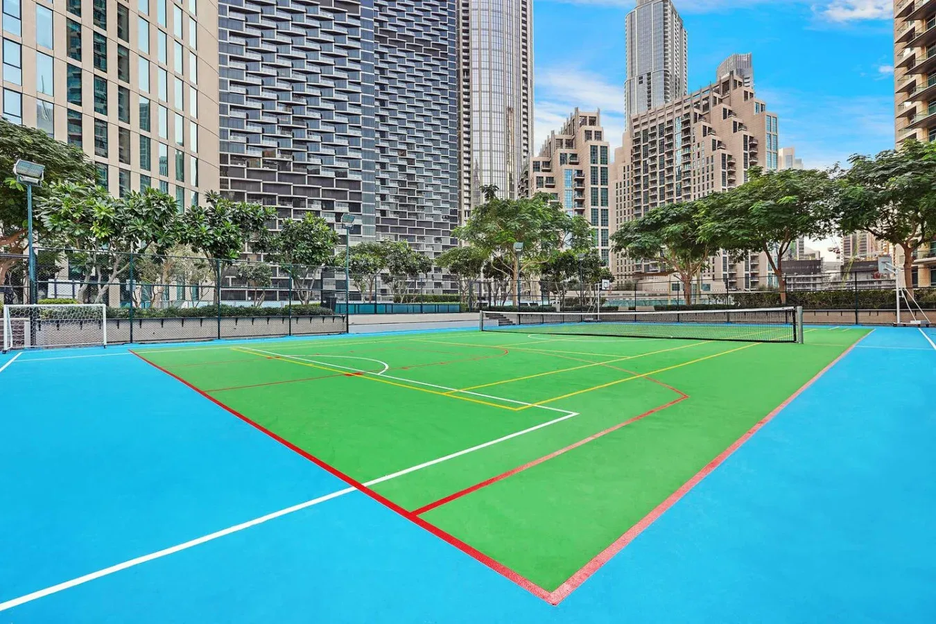 An outdoor sports court has green playing areas, blue borders, and tall buildings nearby.
