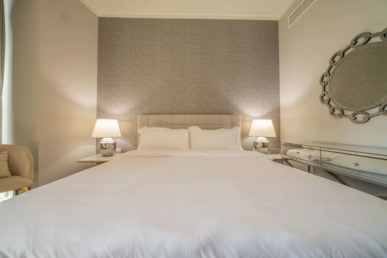 A bedroom with a neatly made bed, bedside lamps, and a decorative wall mirror.