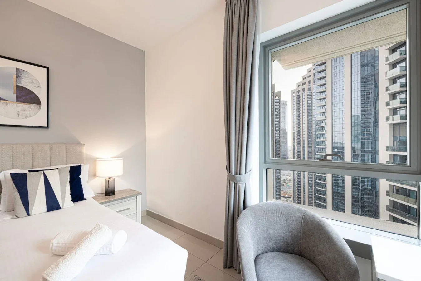 A bedroom with a bed, bedside lamp, and a large window overlooking nearby buildings.