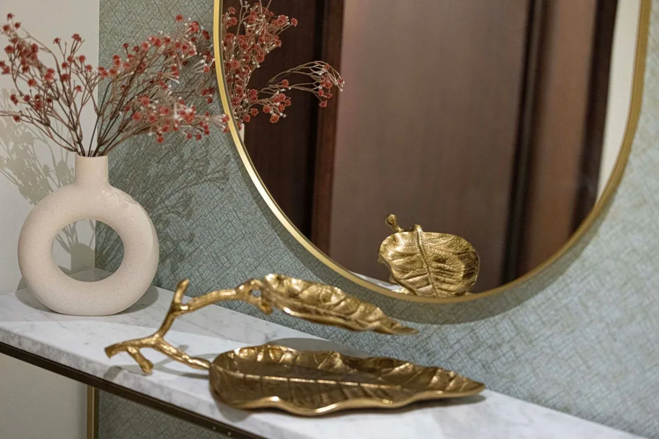 A decorative tabletop with a round mirror, a vase of dried flowers, and gold-toned dishes.