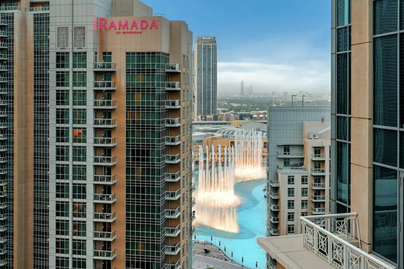 A city skyline overlooks a turquoise pool and illuminated fountains between high-rise buildings.