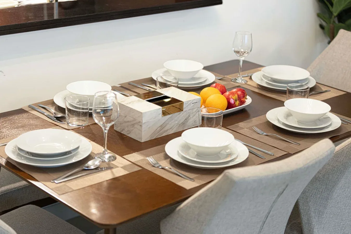 A dining table is set with plates, glasses, and a bowl of fruit.