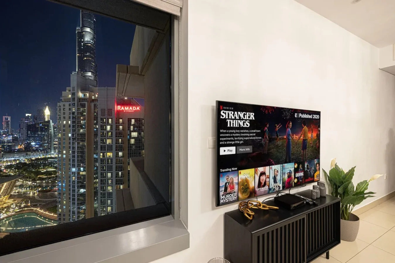 A living area with a television displaying a show beside a large window overlooking city lights.