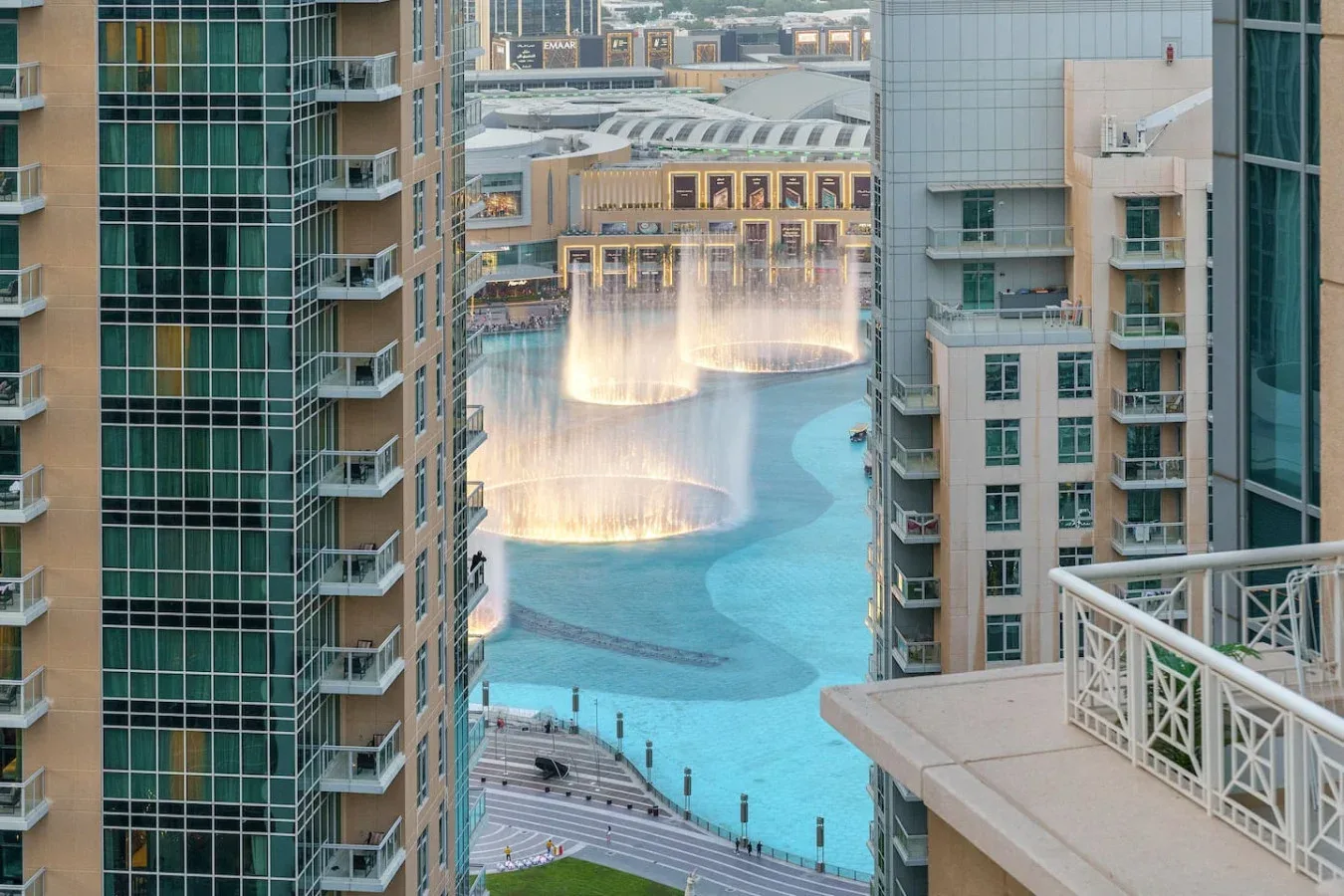 A cityscape overlooks a turquoise pool and illuminated fountains between tall buildings.