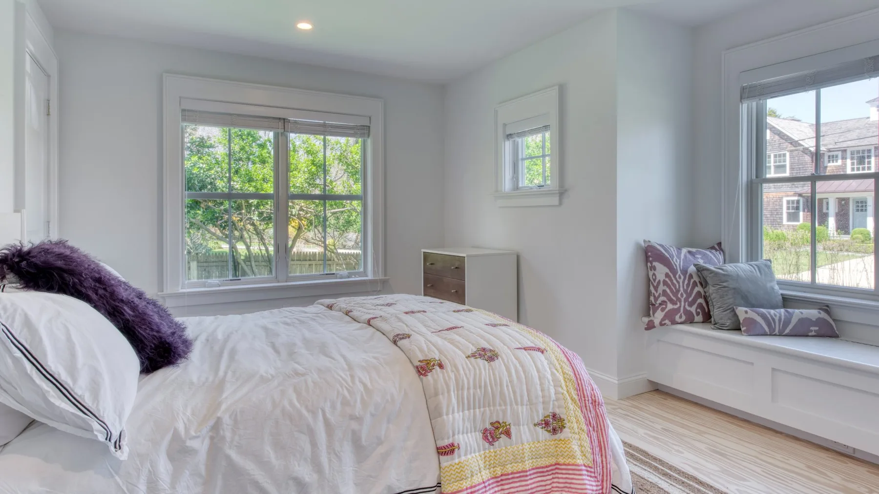 Un dormitorio luminoso con una cama bajo una ventana grande y un asiento junto a otra ventana.