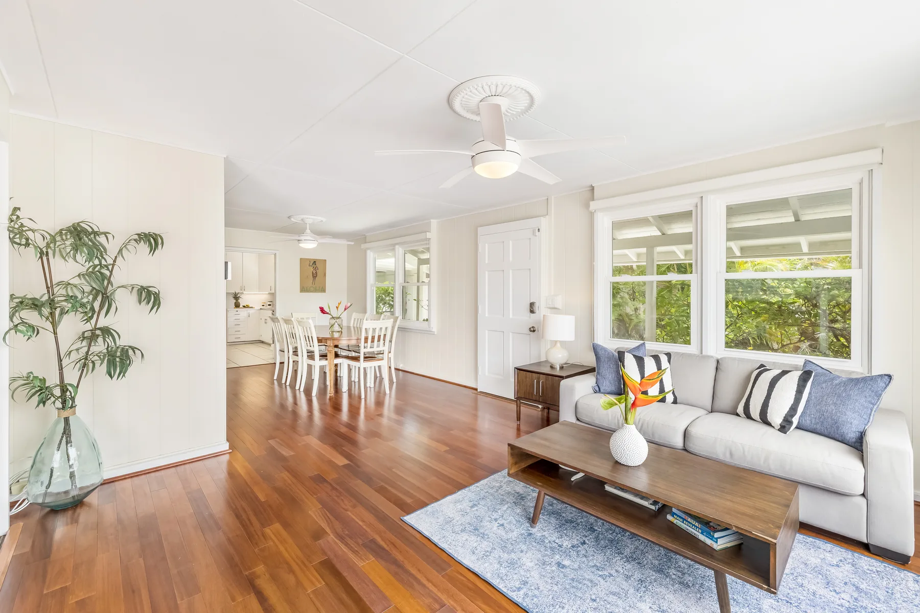 A bright living room opens onto a dining area, with large windows and wood flooring.