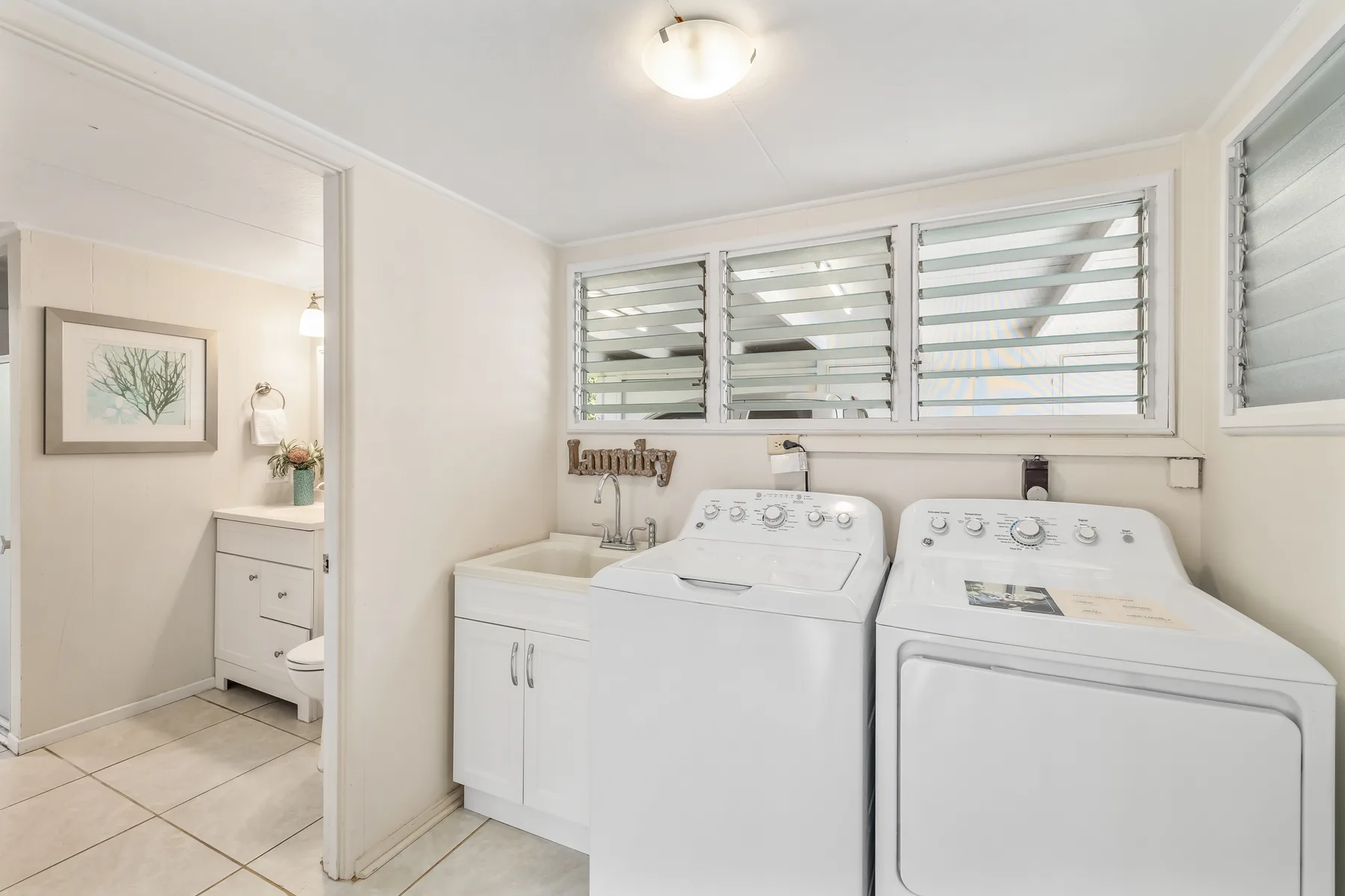 A laundry room with a washer and dryer beneath louvered windows.