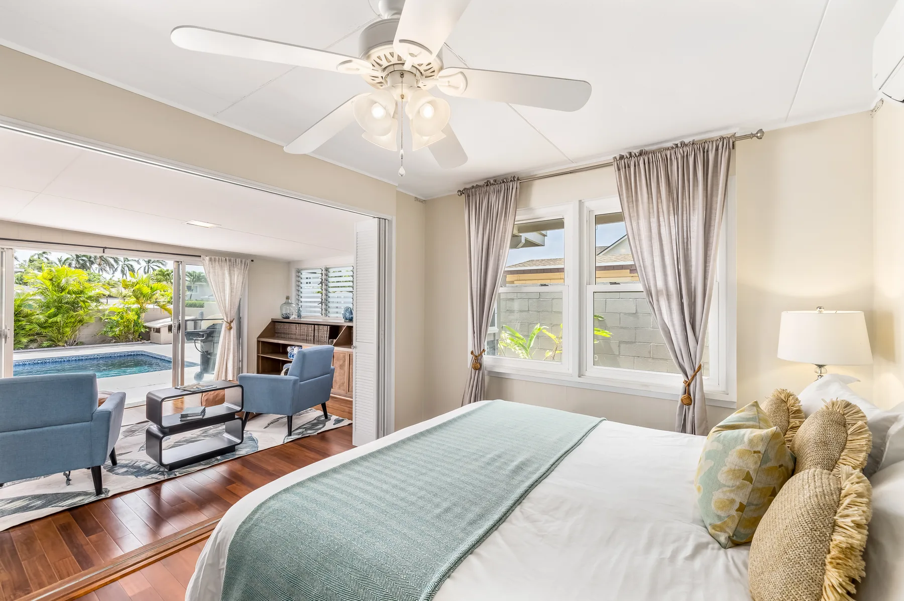 A bedroom with a bed beside windows overlooking a living area and pool.