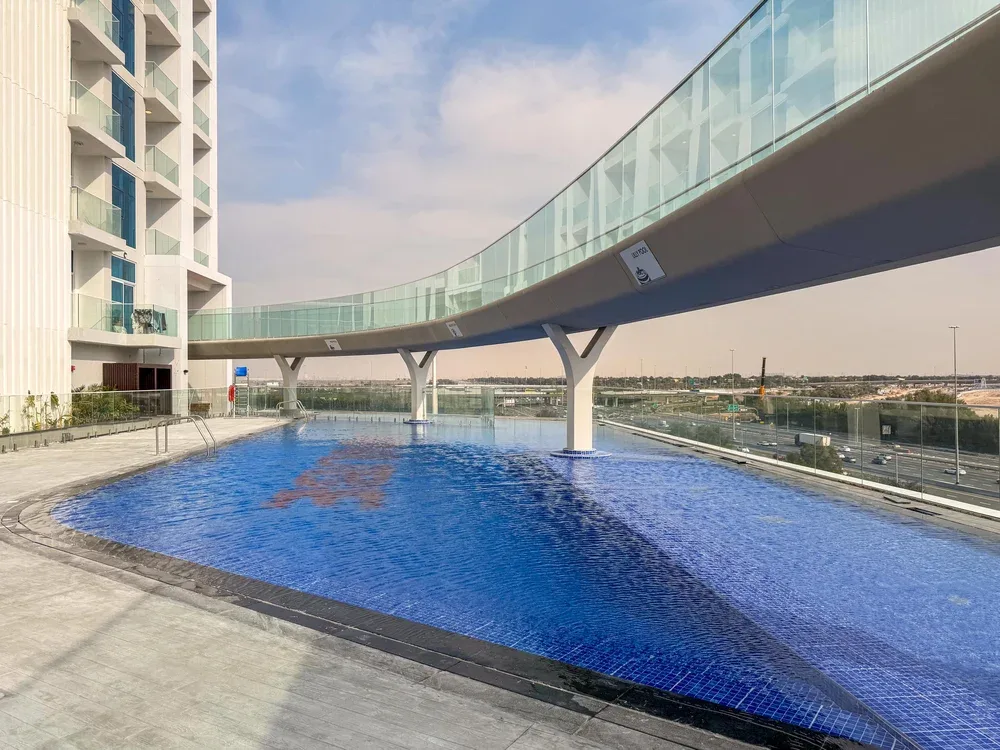 A blue swimming pool sits beside a modern building and curved glass walkway.