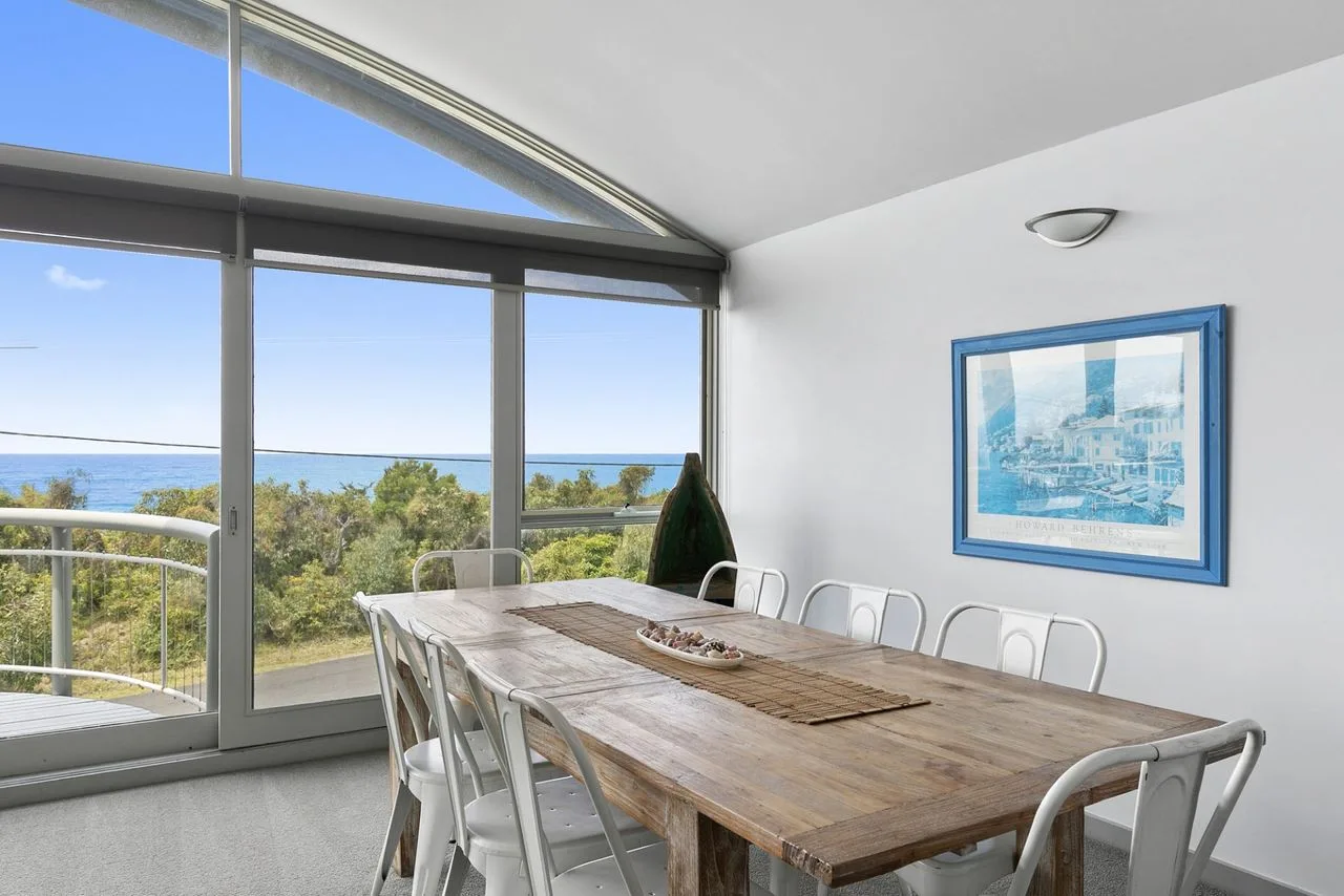 Comedor con una mesa larga junto a grandes ventanas con vistas al agua.