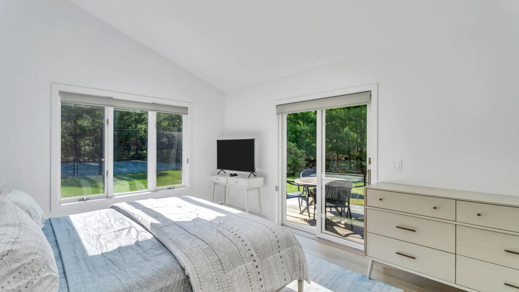 Un dormitorio luminoso con cama, cómoda y ventanas con vistas a la vegetación.