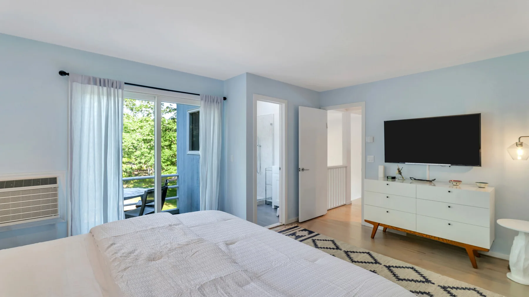 Un dormitorio luminoso con una cama, un televisor y una puerta de vidrio hacia la vegetación.