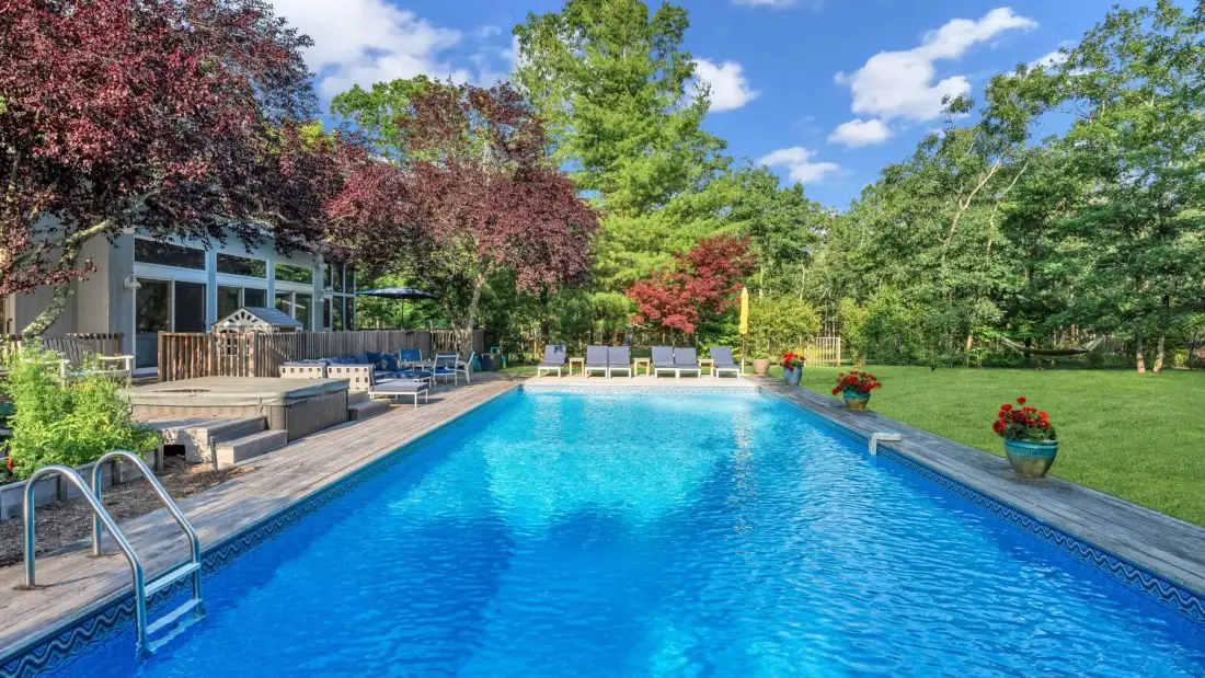 Una piscina junto a una terraza de madera y árboles alrededor.
