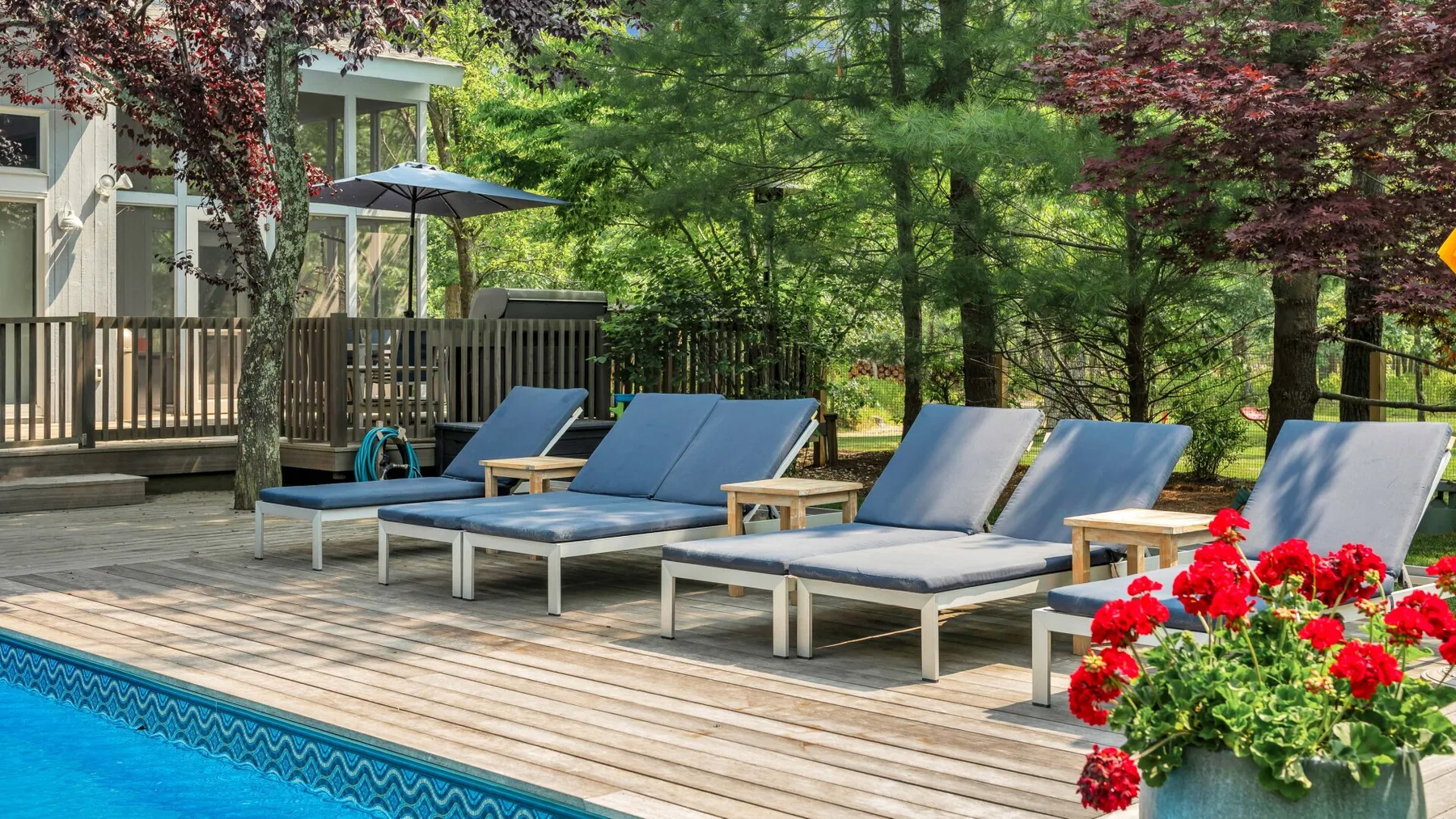 Una piscina exterior junto a una terraza de madera con tumbonas y flores rojas.