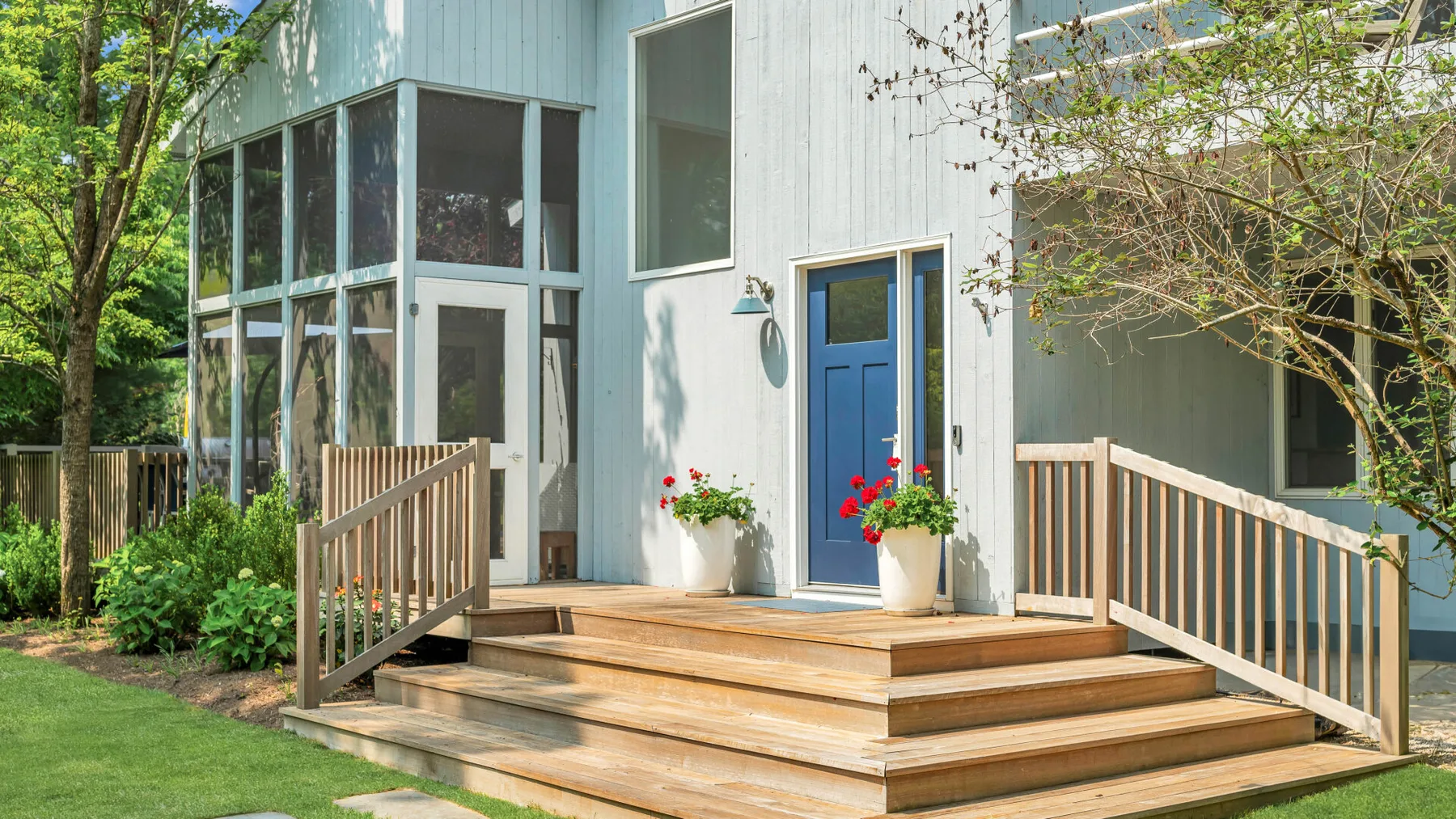 Una casa de color claro con puerta azul da a una terraza de madera rodeada de vegetación.