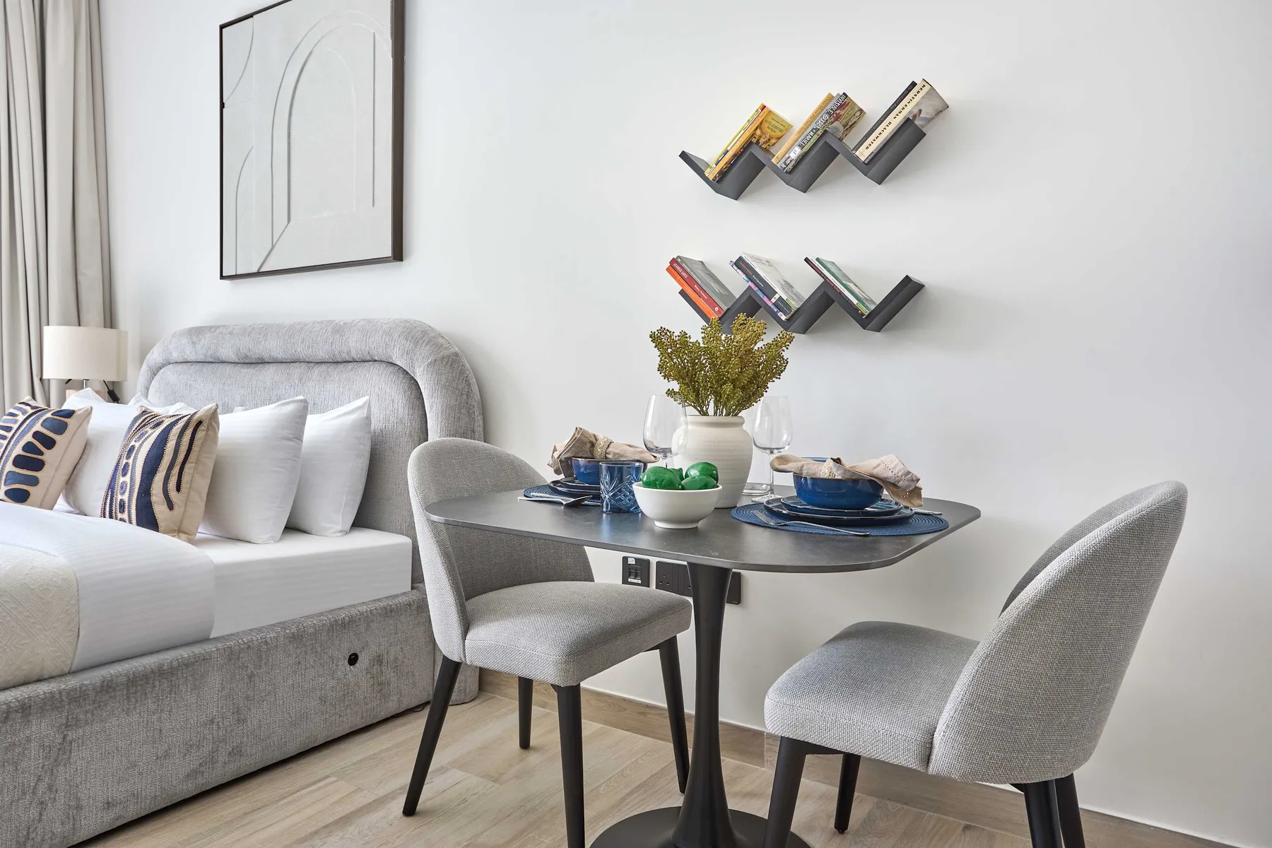A bright bedroom with a bed, a small dining table, and wall-mounted shelves.