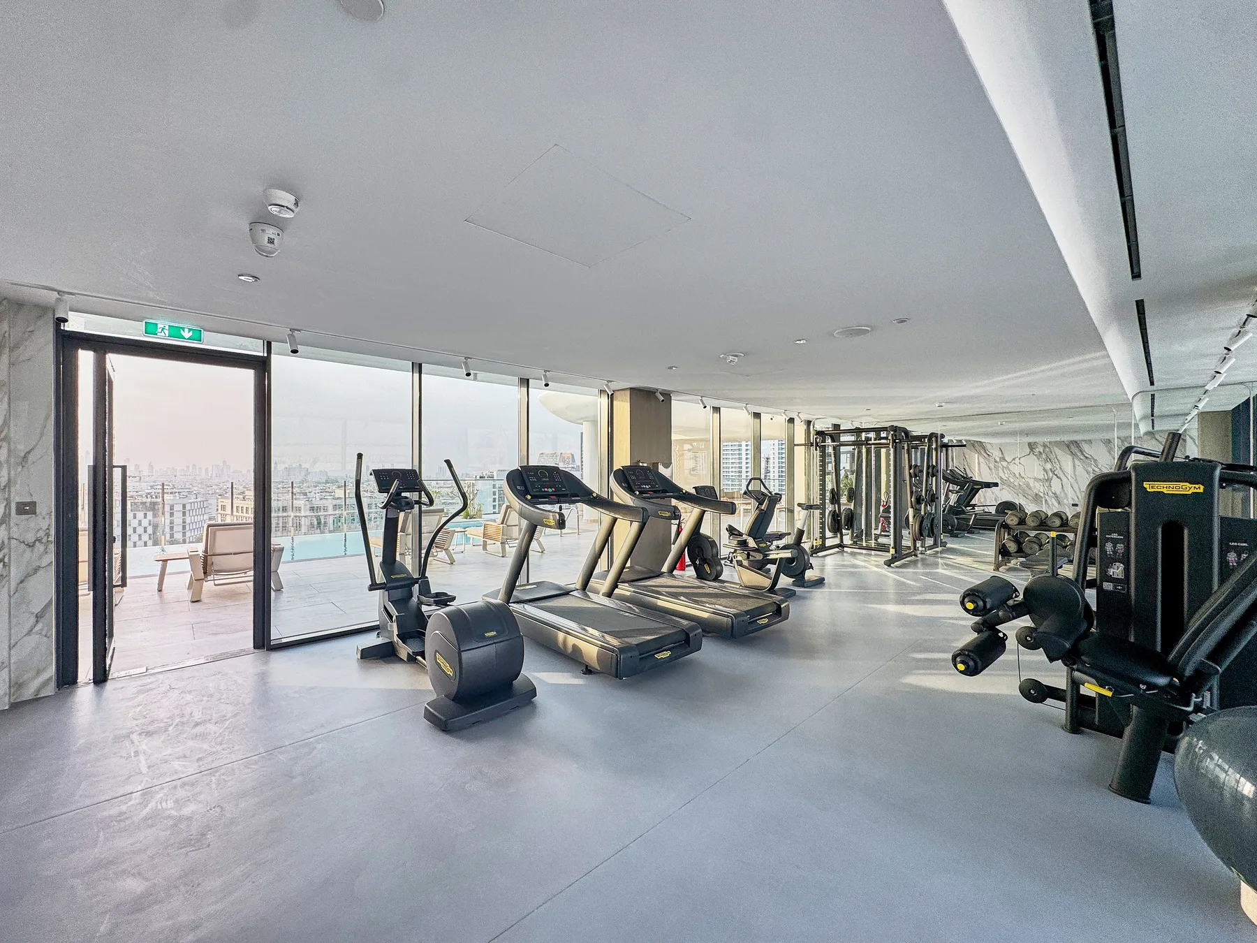 A bright gym with treadmills and strength-training equipment beside large windows.