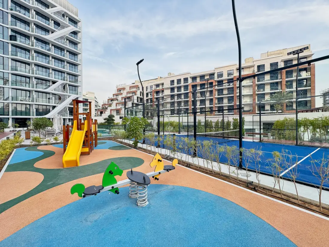 A colorful outdoor playground with a slide and spring riders beside a fenced court.