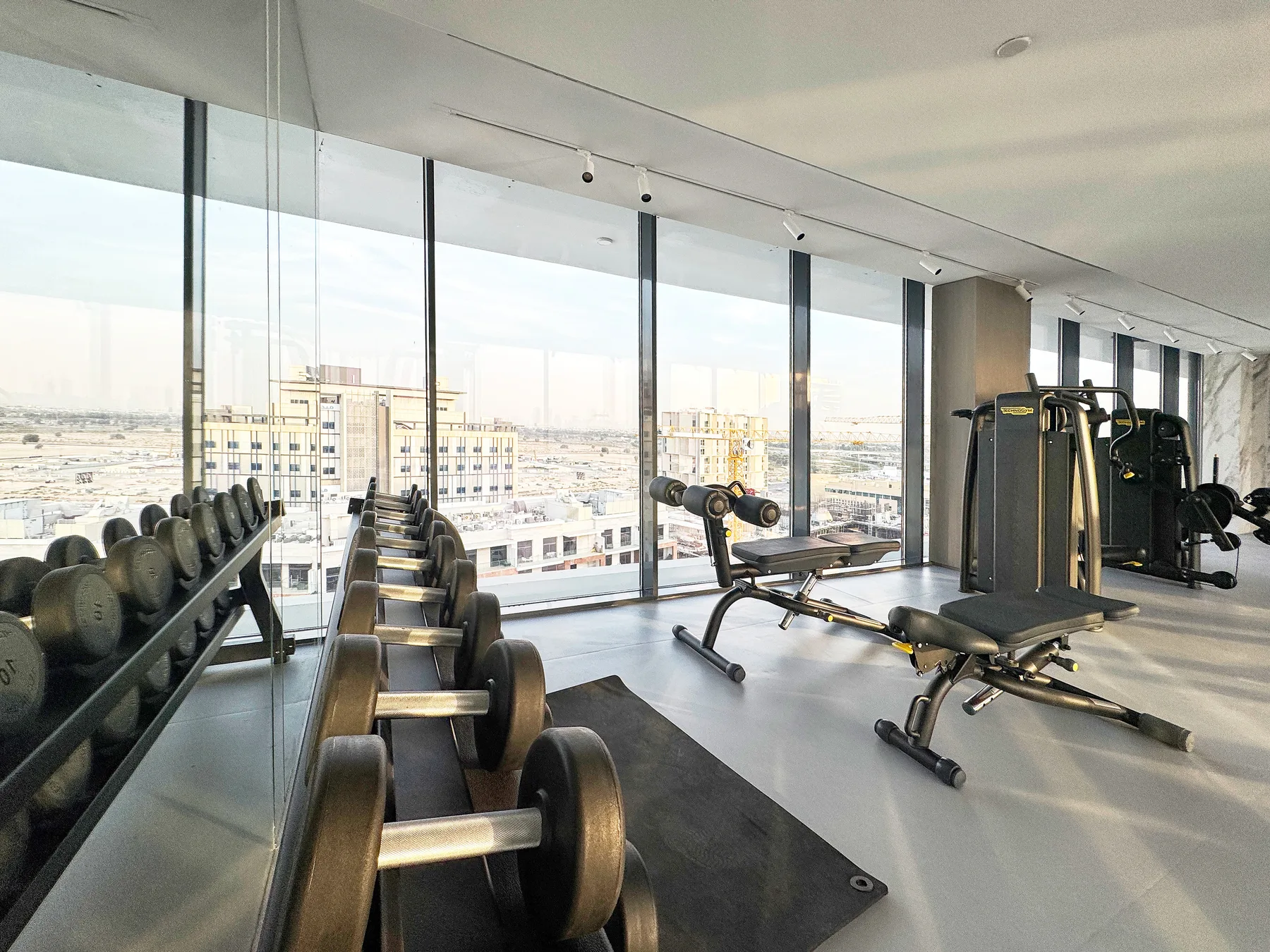 A gym with weight benches and exercise machines beside floor-to-ceiling windows.
