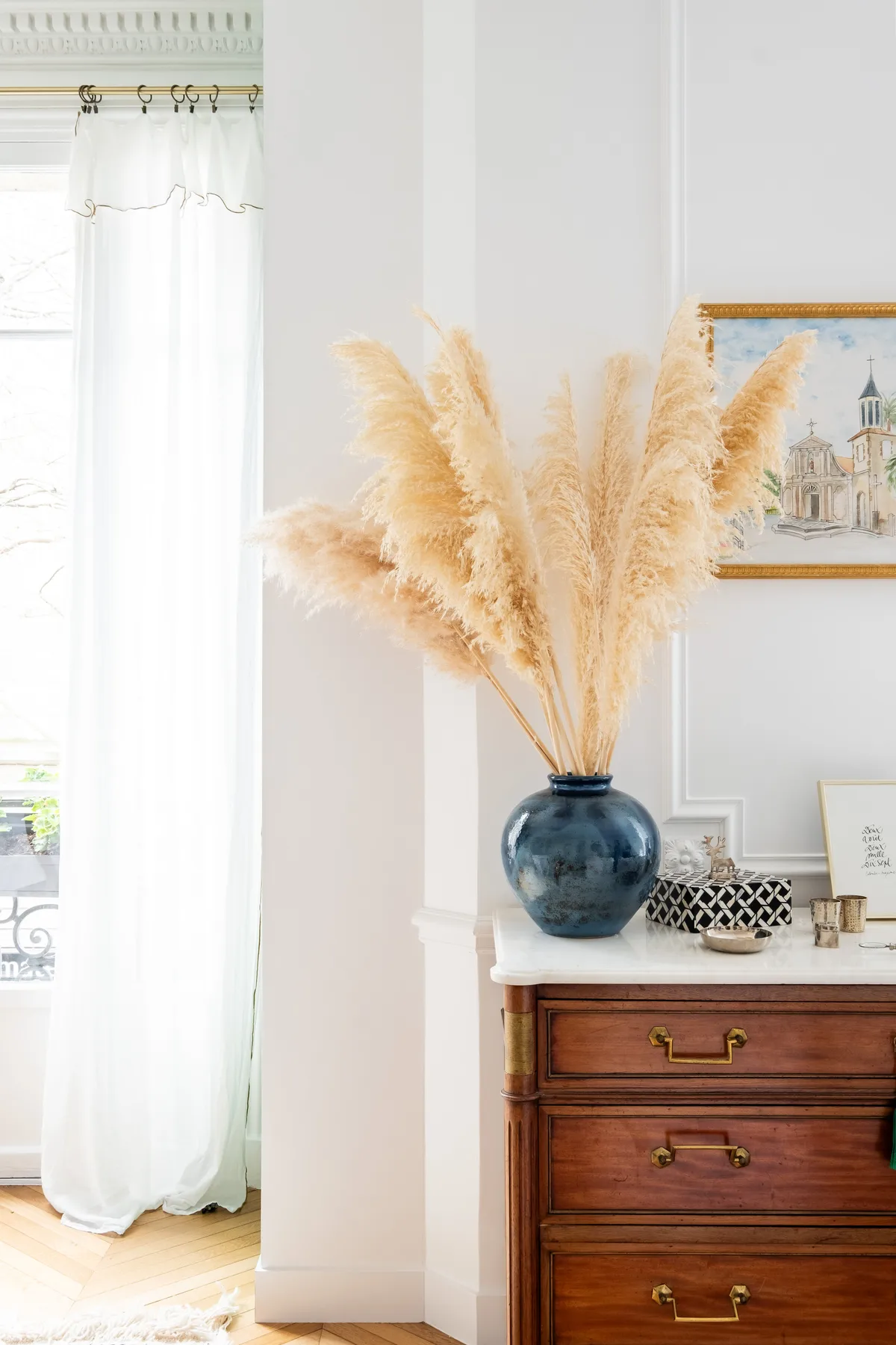 A wooden dresser with a blue vase of dried grasses stands beside a curtained window.
