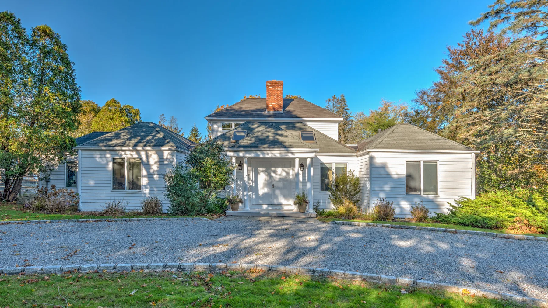 A white house stands beside a gravel driveway, surrounded by trees.