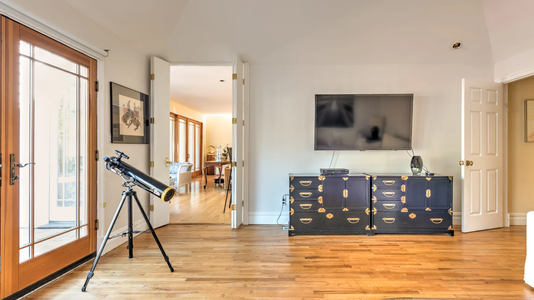 A bright bedroom with a telescope beside a dresser and television.