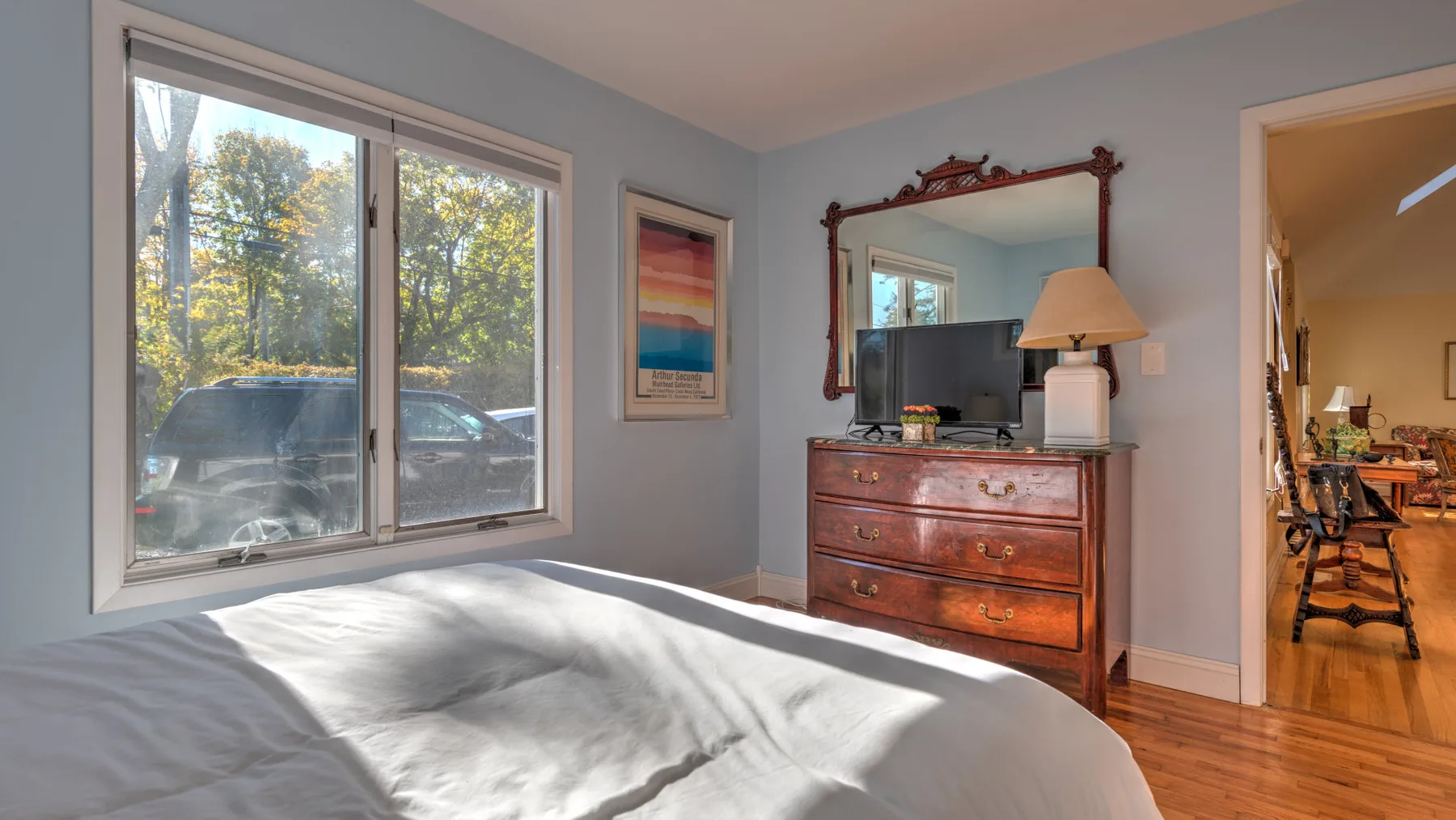A bedroom with a bed beside a large window overlooking trees and parked cars.
