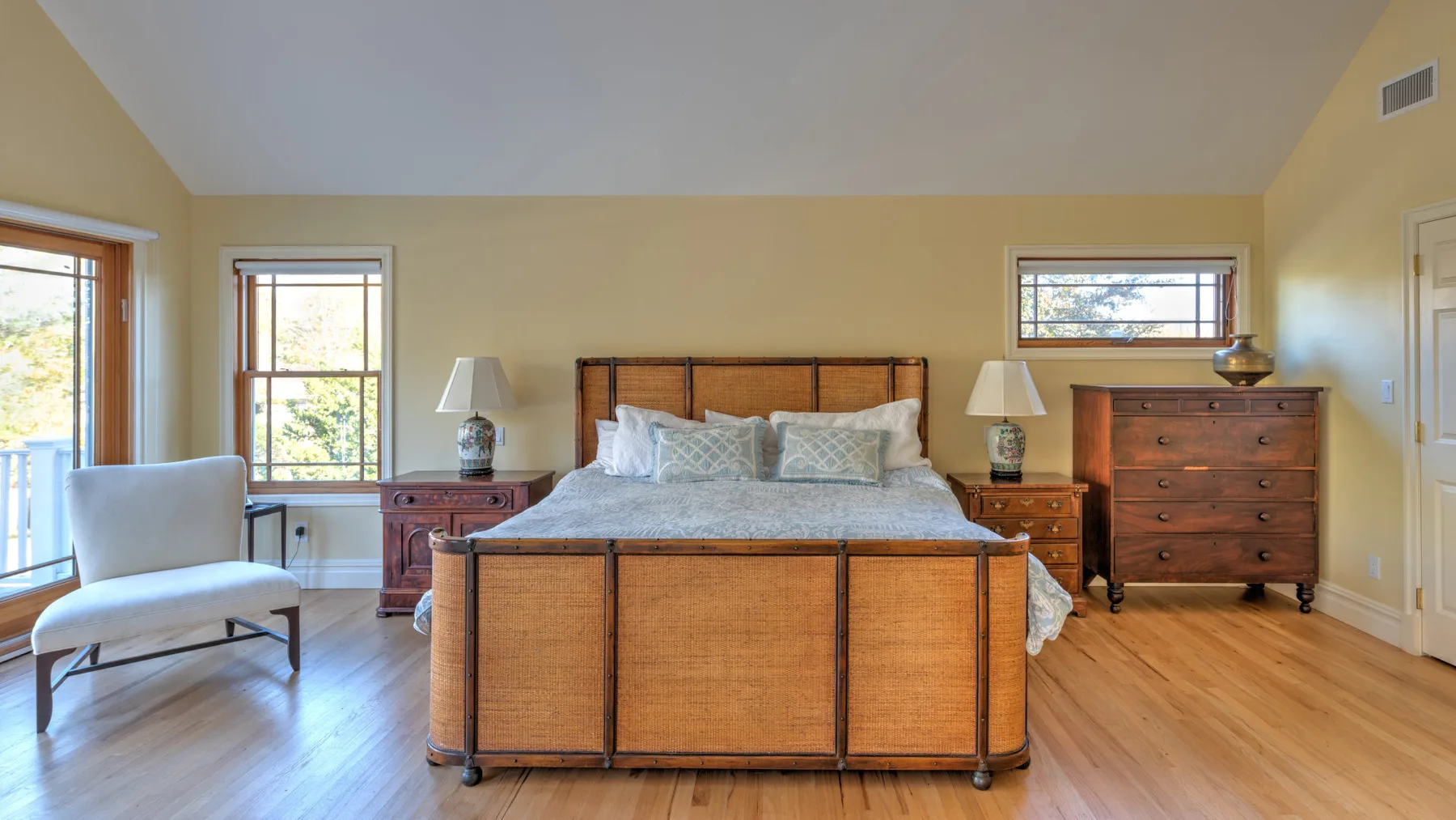 A bedroom with a large bed, wooden dressers, and a chair beside the windows.