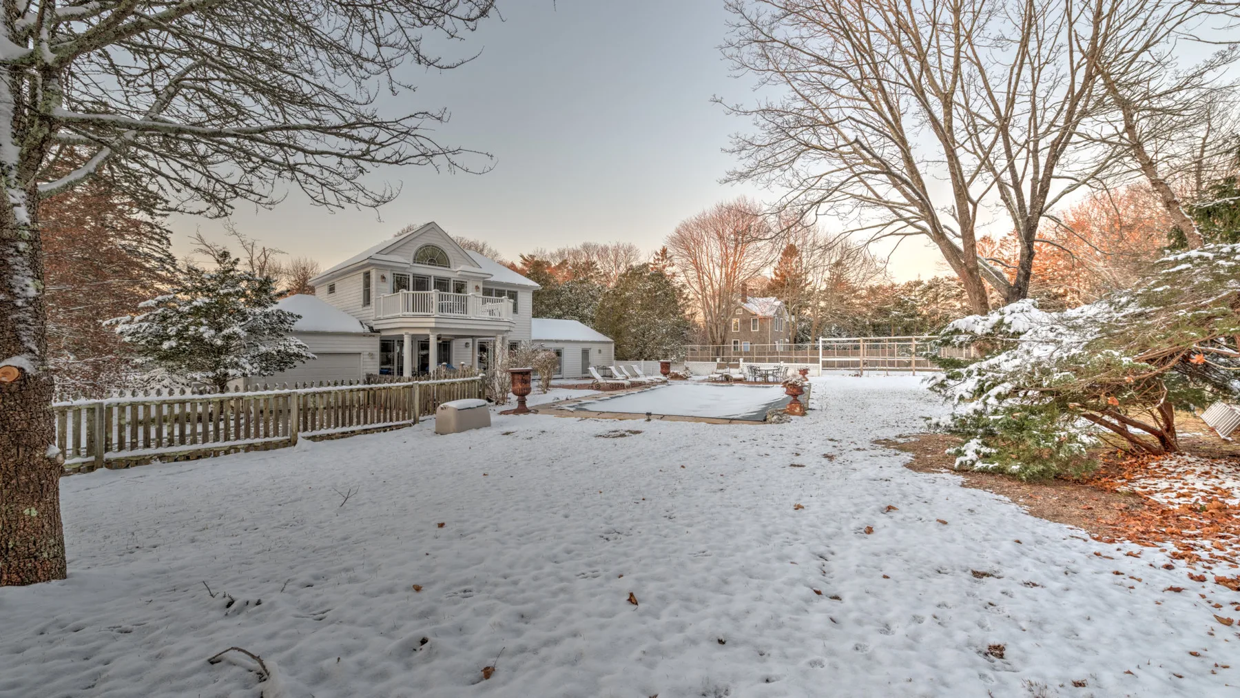 A snow-covered backyard with a large house, bare trees, and a fenced pool area.