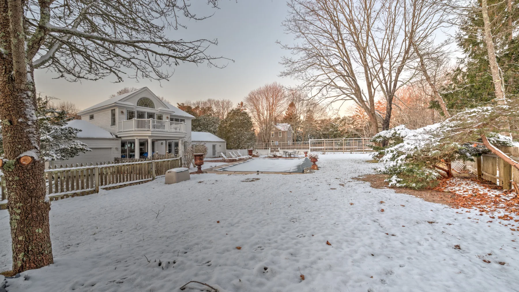 A snow-covered yard sits in front of a large house and bare trees.