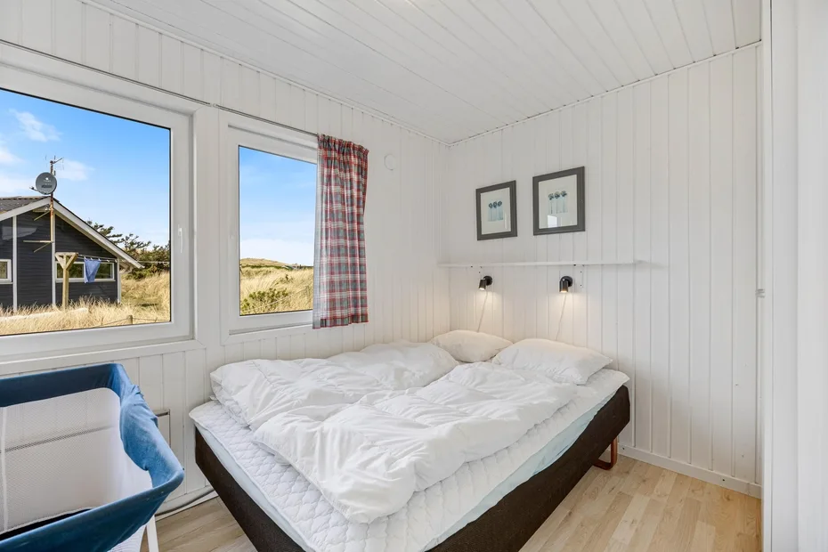Un dormitorio con una cama bajo dos cuadros enmarcados y ventanas con vistas al exterior.