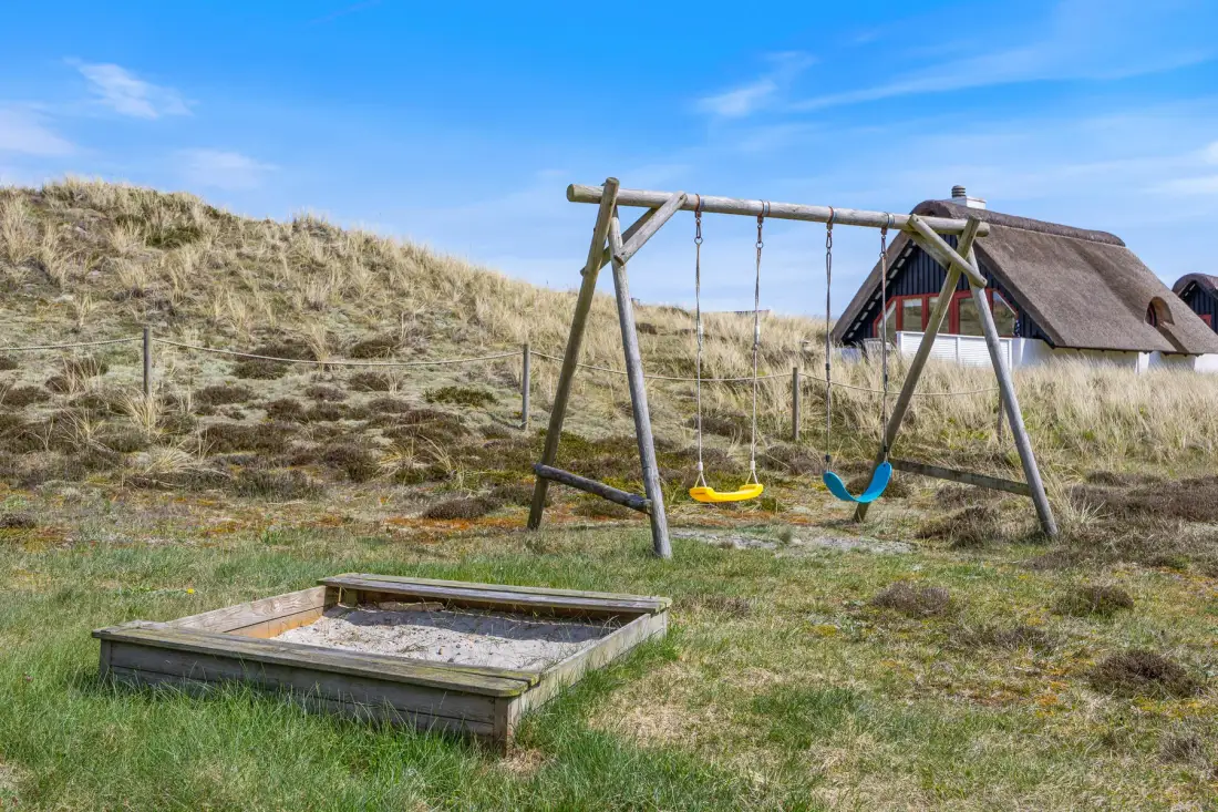 Un jardín con columpios de madera y un arenero junto a una casa con techo de paja.