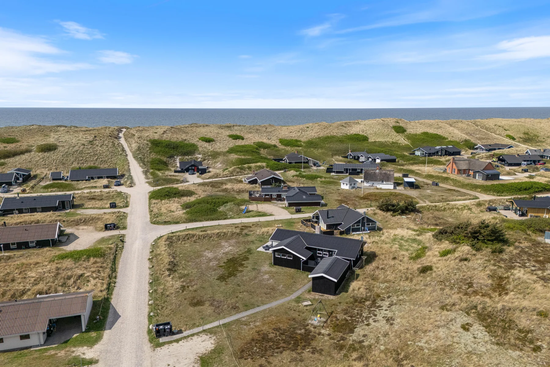 Una vista aérea muestra casas junto a un camino costero de arena, dunas cubiertas de hierba y el mar.