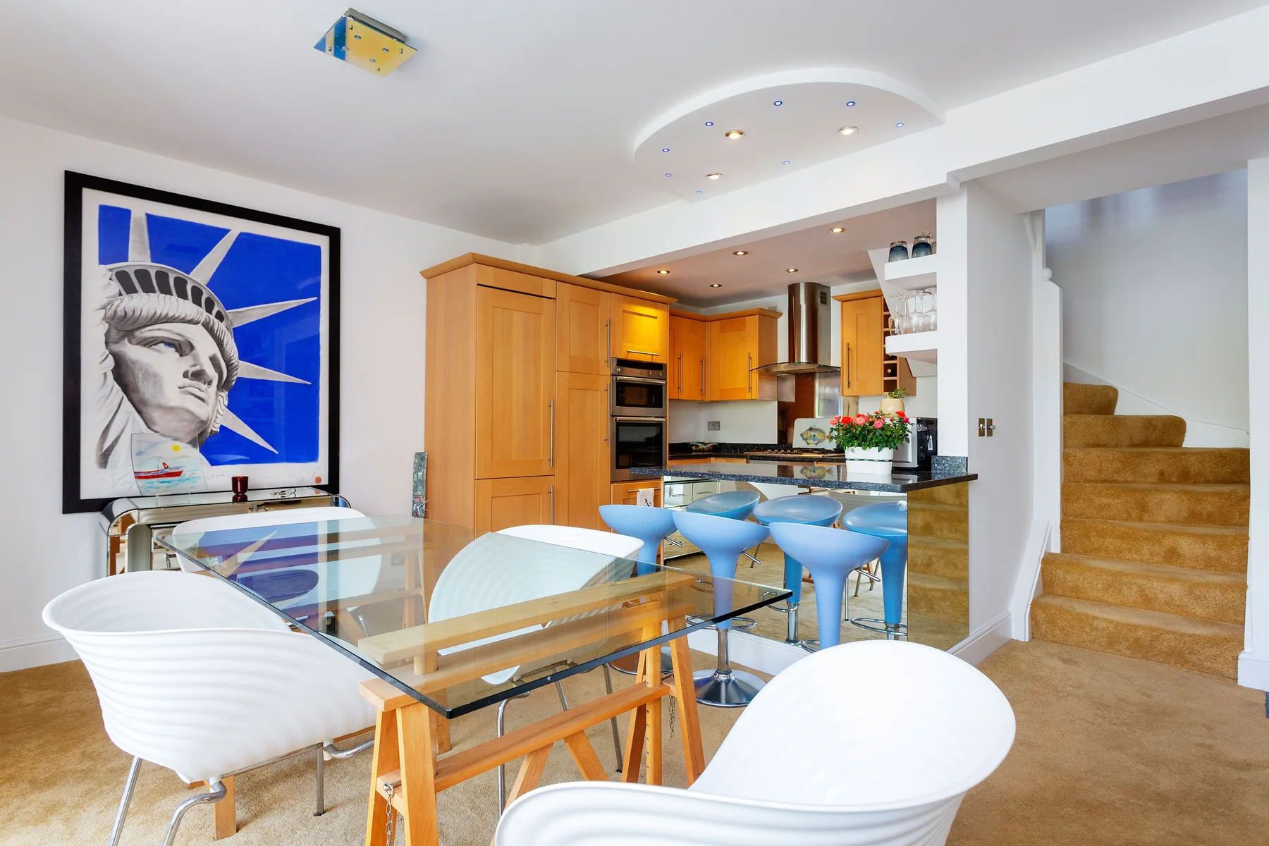 A dining area with white seating and a glass table beside a wood-toned kitchen.