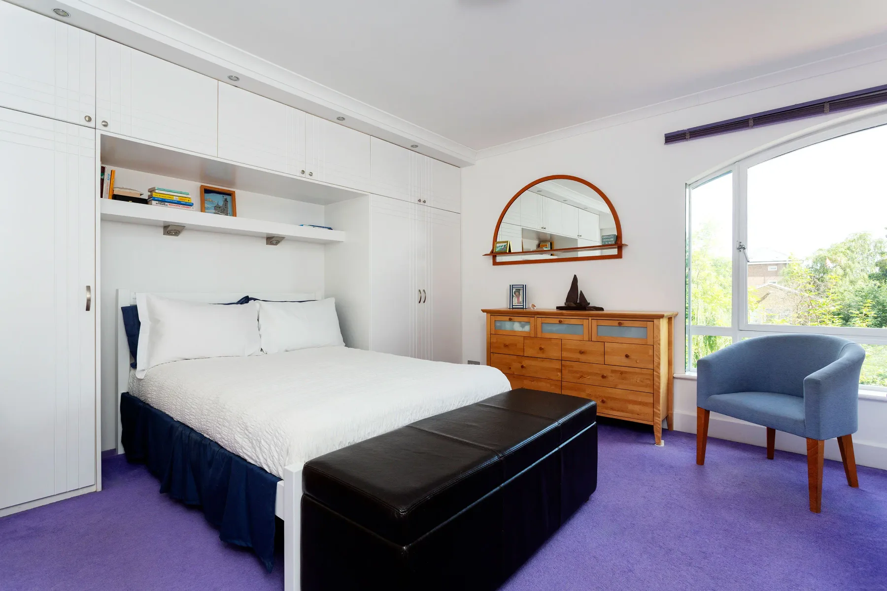 Dormitorio con cama, almacenamiento integrado y una silla azul junto a la ventana.