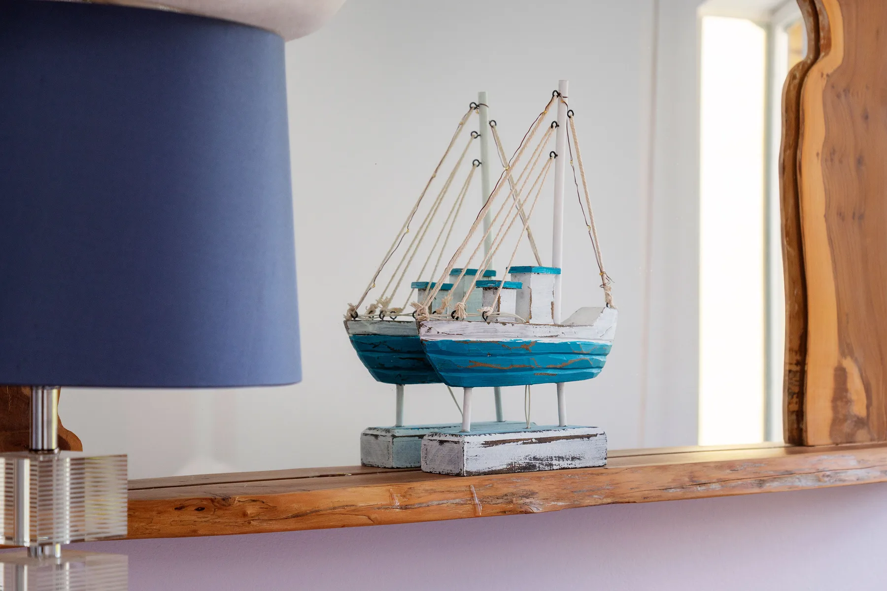 Un barco decorativo en miniatura está sobre un estante de madera junto a una lámpara azul.